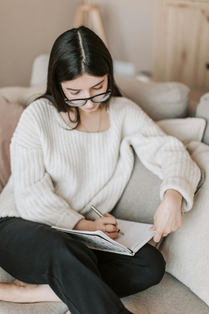 A young woman writes in a notebook while sitting comfortably on the sofa at home.