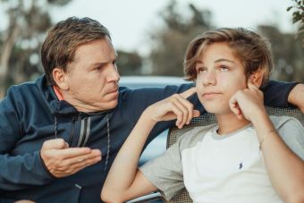 Father expressing frustration while teenage son shows defiance, seated outdoors.