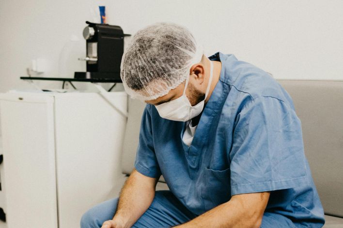 A male doctor in hospital attire sitting pensively, representing healthcare challenges.